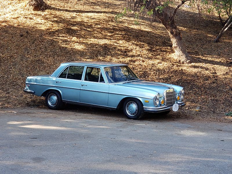 Classic blue vintage car parked beside a dirt slope with sparse trees.