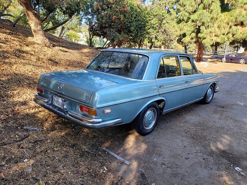 Vintage blue sedan parked on a dirt path under trees.