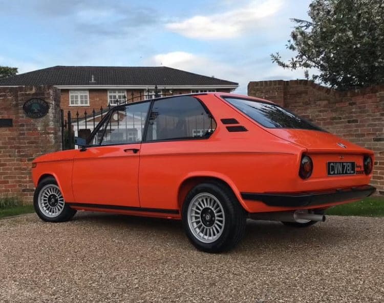Bright orange classic car parked outside a house on a cloudy day.