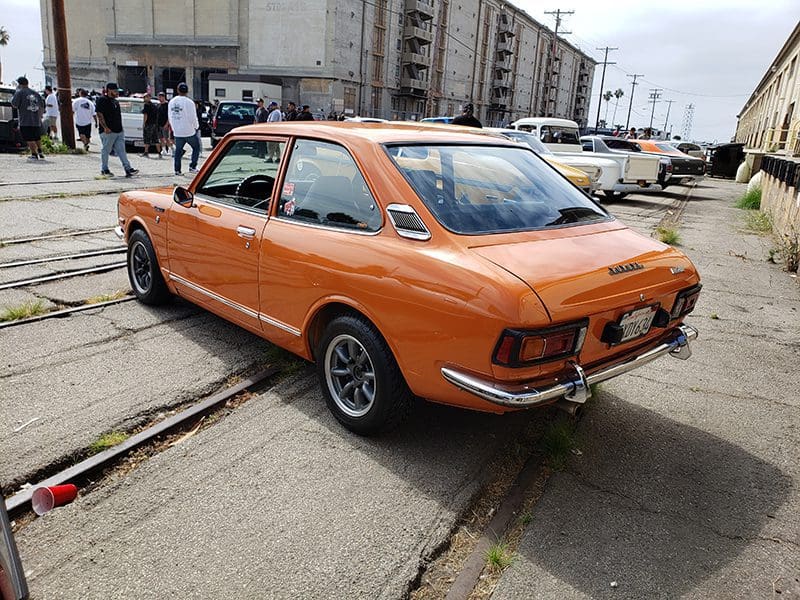 Orange vintage car parked near an industrial building on a cloudy day.