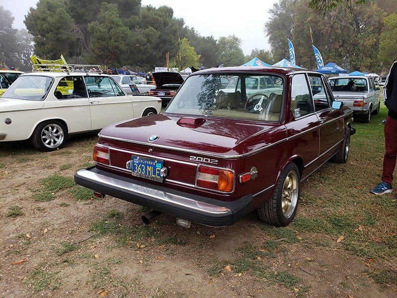 A classic maroon car parked on grass with other vintage cars around.