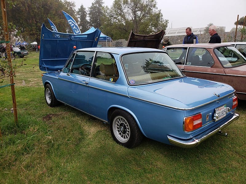 Classic blue BMW 2002 with open hood and trunk at a car show.