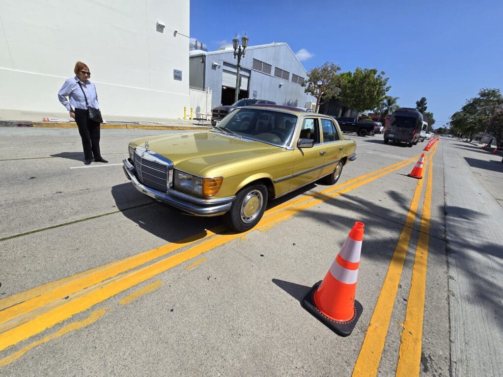 A vintage gold car parked beside orange cones on a sunny street.