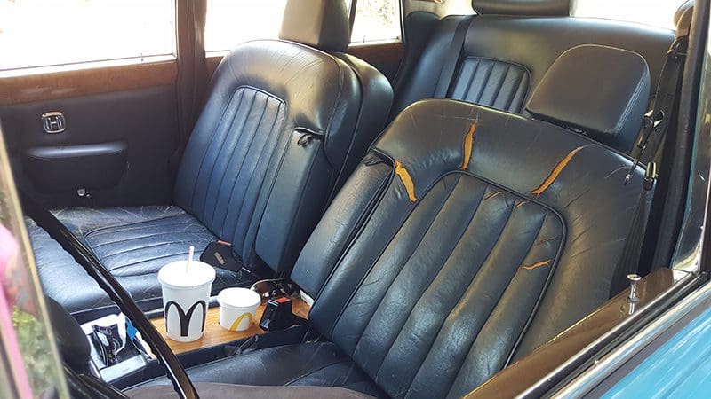 Vintage car interior with black leather seats and a wooden tray holding a drink.
