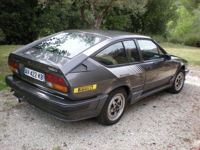 Black classic Opel Calibra parked on a gravel path surrounded by greenery.