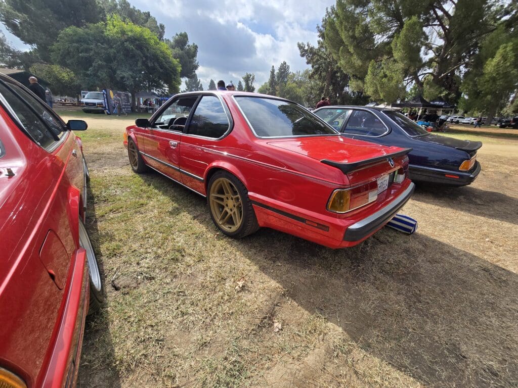 Red vintage BMW parked on grass with bronze wheels under cloudy sky.