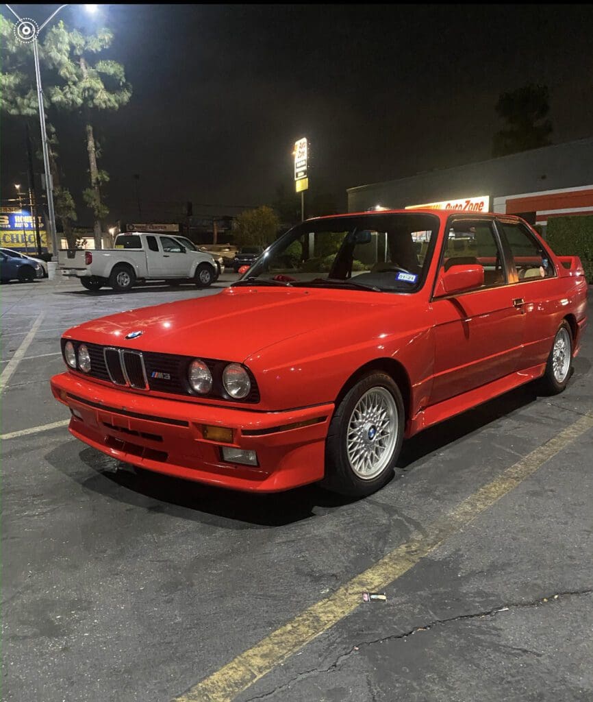 Classic red BMW E30 parked in a nighttime lot.