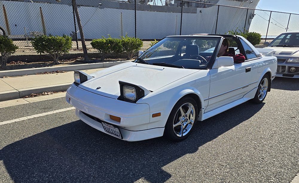 White convertible sports car with pop-up headlights in a sunny parking lot.