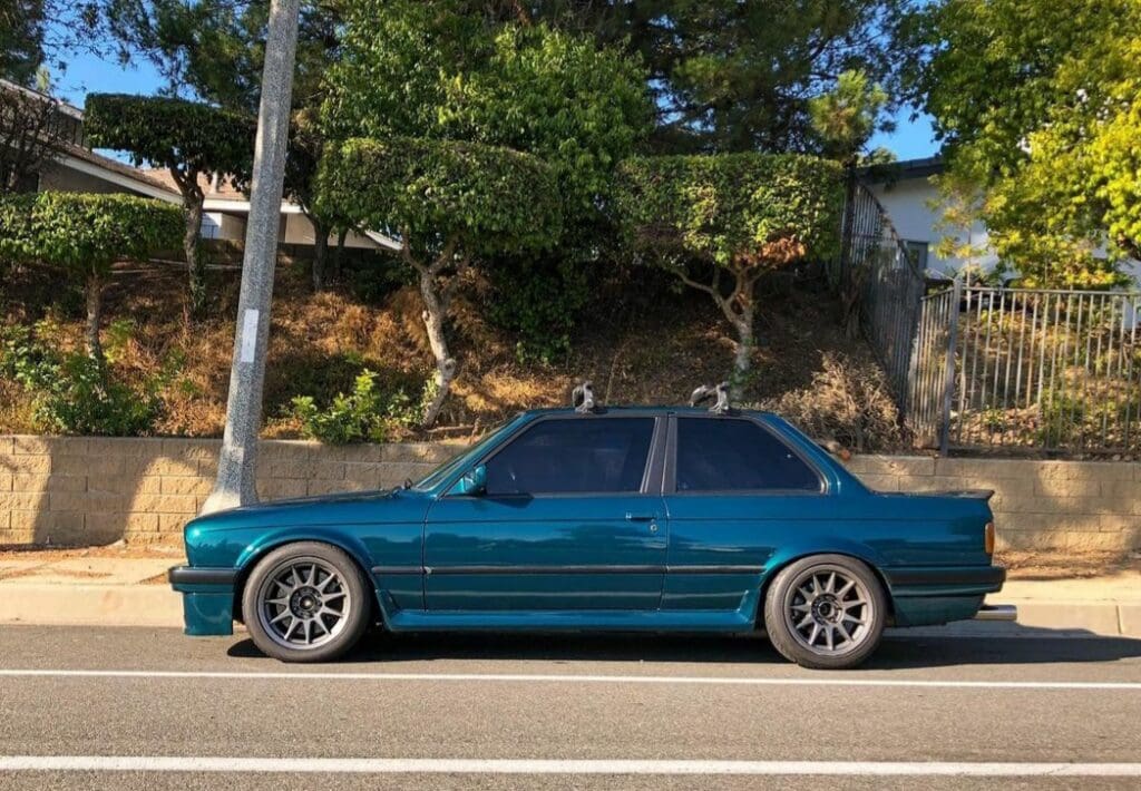 A teal sedan parked on the street near trees and a rocky hill.