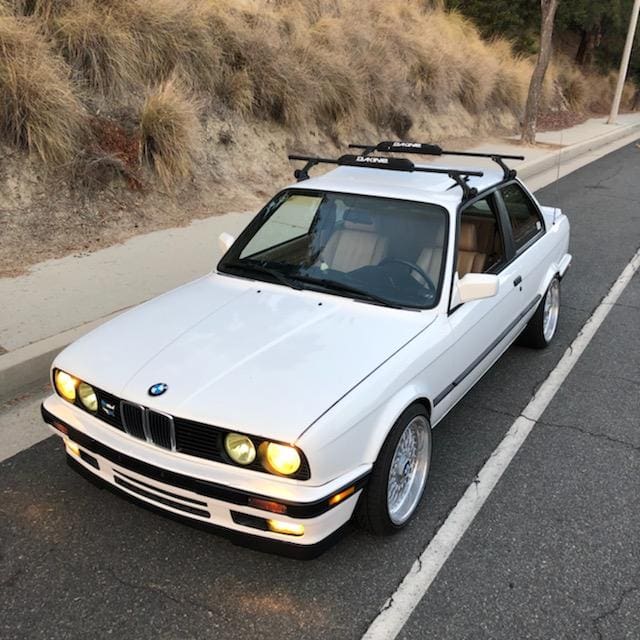 Classic white BMW E30 with roof rack parked on a roadside.