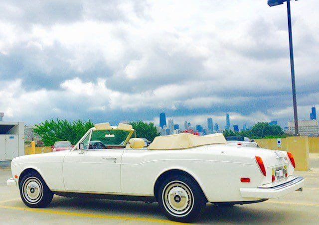 Classic white convertible car parked with a city skyline backdrop under a cloudy sky.