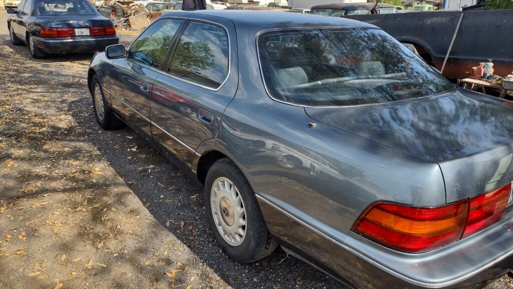 A gray vintage sedan parked on a dirt surface with trees reflected on its windows.