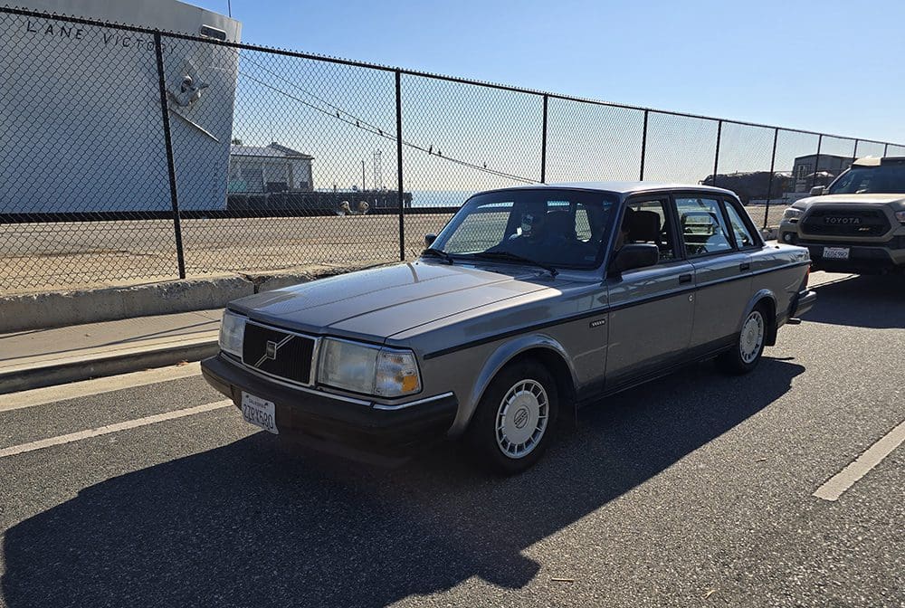 Classic silver Volvo sedan parked by a fenced area under clear blue sky.