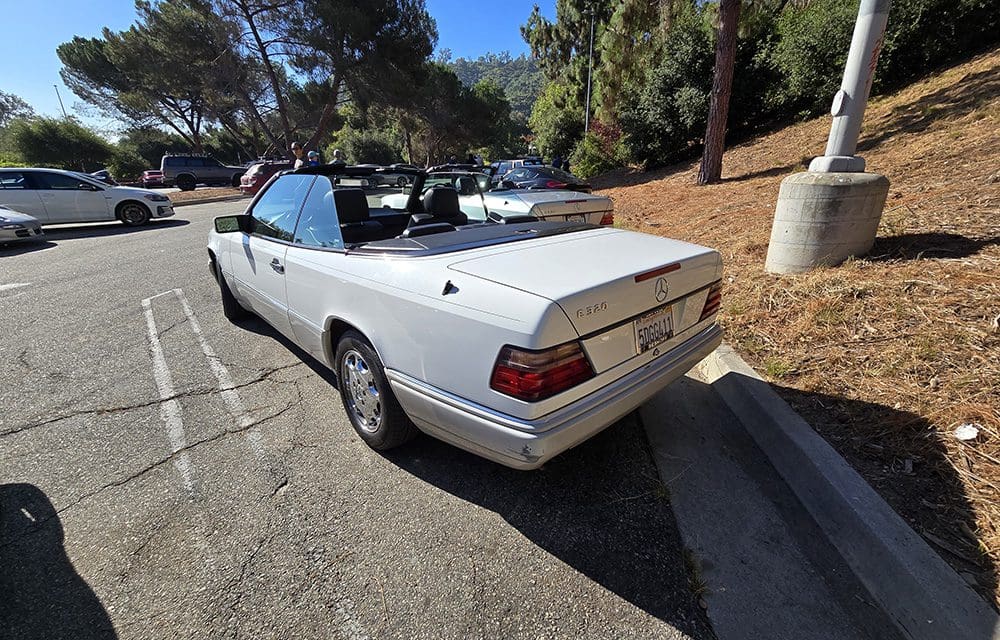 White convertible car parked in a sunny lot with trees around.