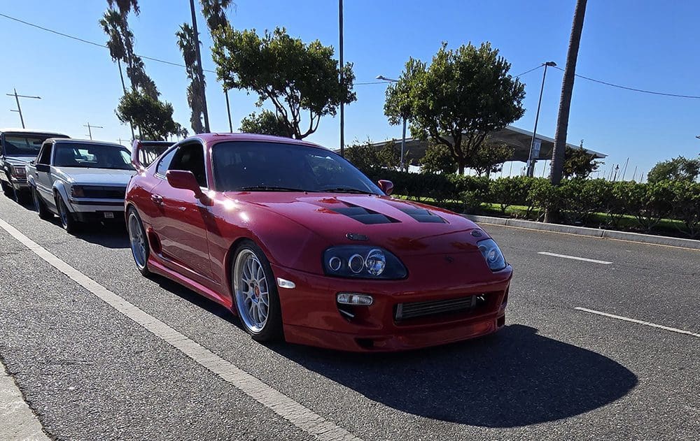 Red sports car parked under trees in a sunny lot.