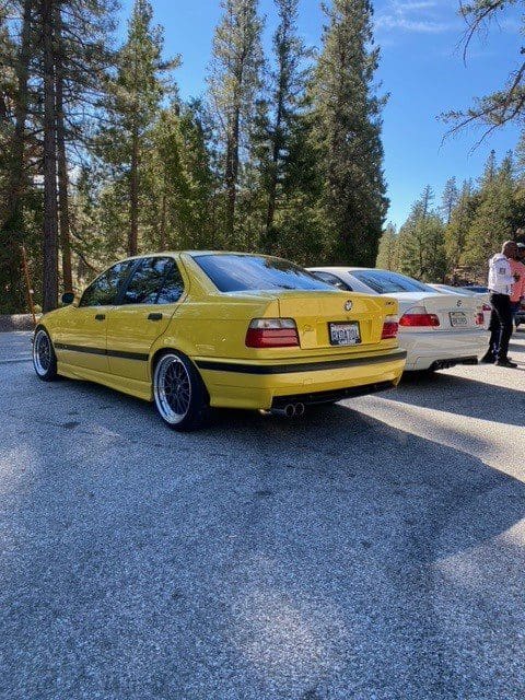 Yellow BMW E30 parked on a sunny street with trees.
