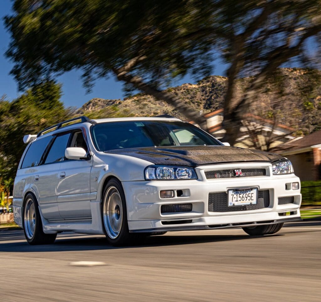 White Nissan Skyline R34 GT-R driving on a road with a tree in the background.
