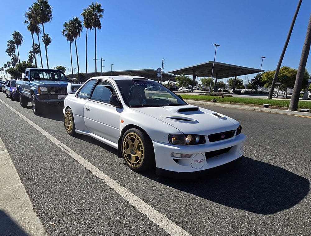 White Subaru Impreza with gold wheels parked near palm trees on a sunny day.