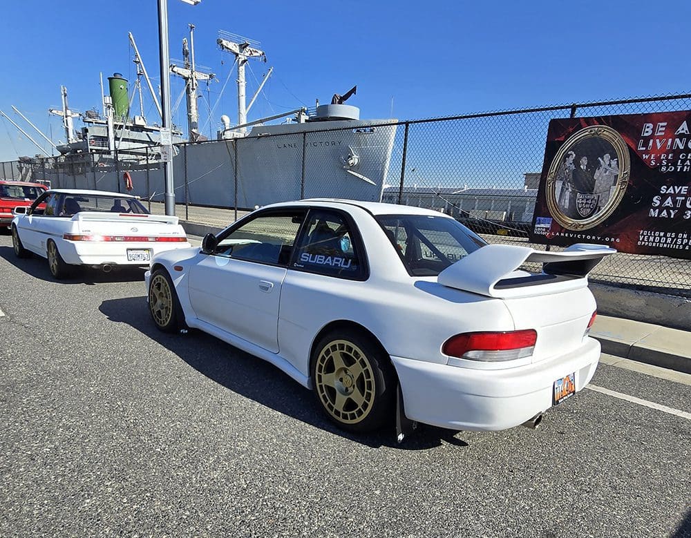 White sports car with gold rims parked near boats on a sunny day.
